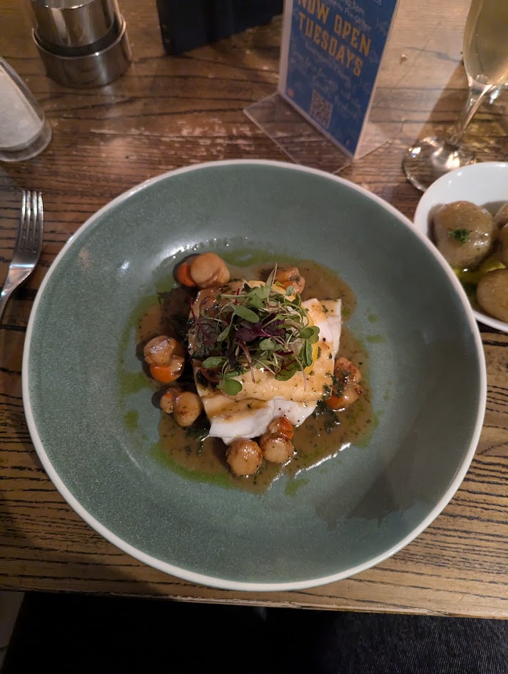 White fish on a blue/grey plate served with a brown butter sauce. A bowl of new potatoes sits beside the dish, as well as a glass of champagne.