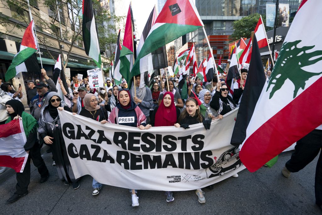 A crowd with hijabi women at the front with a banner that says "GAZA RESISTS, GAZA REMAINS". Around them are Palestine and Lebanon flags. A crowd with hijabi women at the front with a banner that says "GAZA RESISTS, GAZA REMAINS". Around them are Palestine and Lebanon flags.