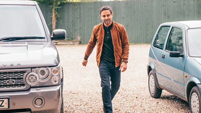 A man walking between two cars - one is an expensive car the other is not.