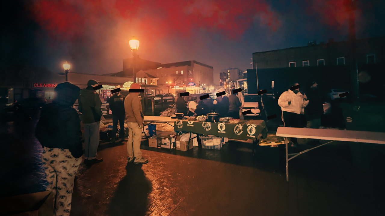 People gathered around a food distribution table at night, with a city street illuminated by lamps in the background.
