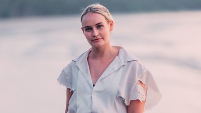 A woman in a white blouse with distressed sleeves stands on a beach