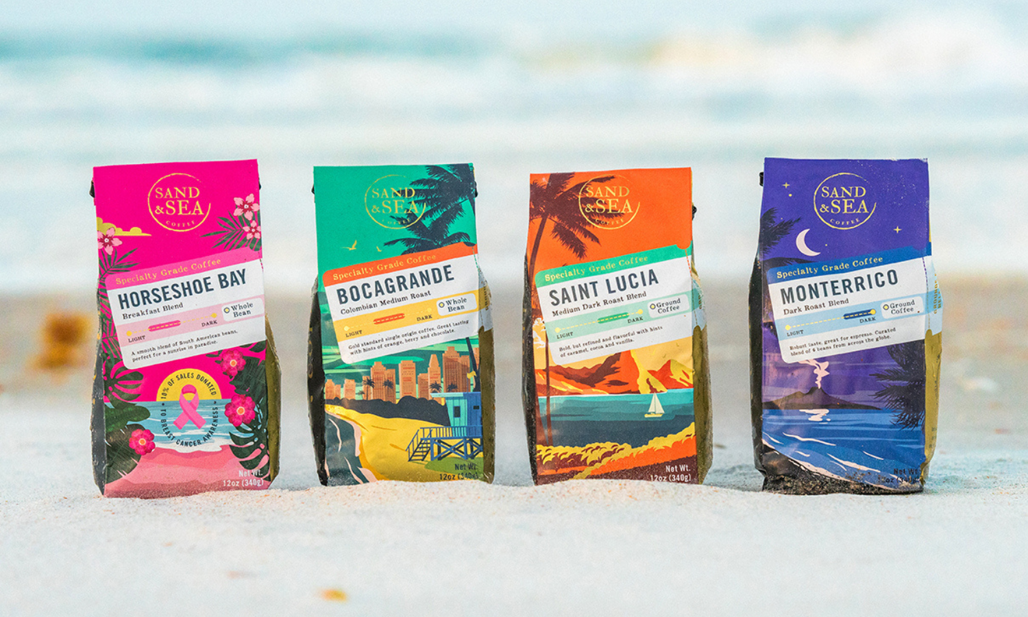 Four colorfully decorated coffee bags rest on the sand with the surf blurred in the background. The bags from left are predominantly pink, green, orange, and purple.