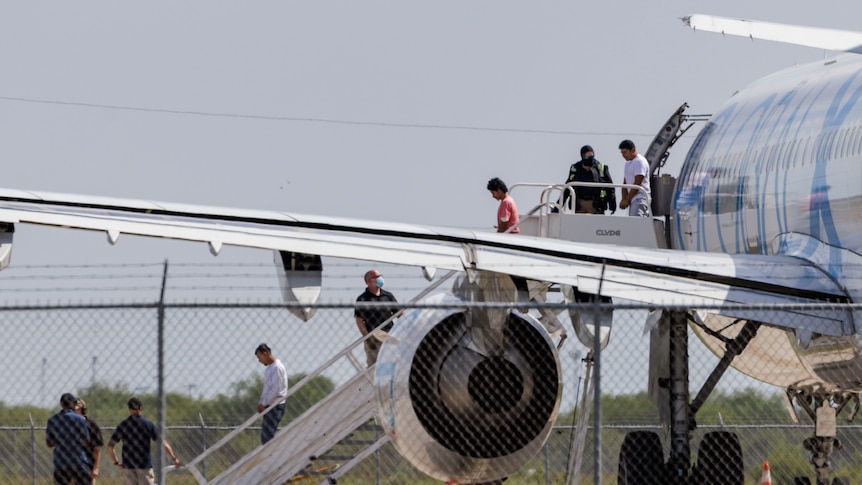 Shackled migrants deplane an aircraft as officials herd them down the stairs.