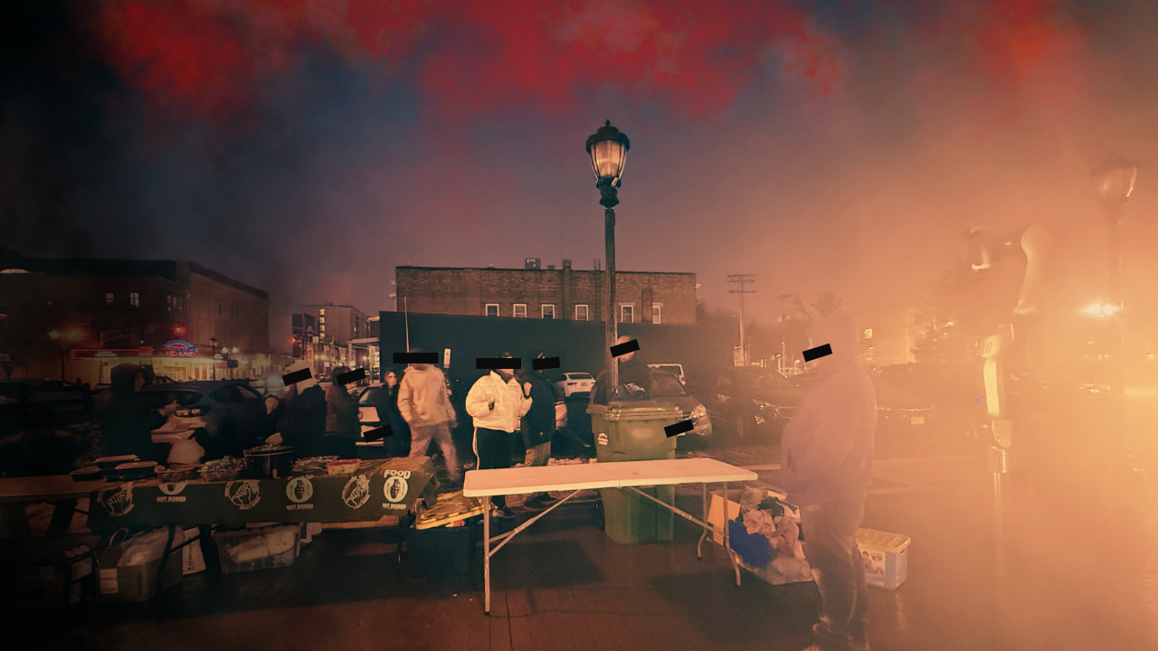 A nighttime scene of a community gathering at a street market, with people milling around tables filled with food and supplies. Some individuals are standing near a green trash bin and a lamppost, while others are engaged in conversation. The atmosphere appears misty or smoky, creating a subdued ambiance.