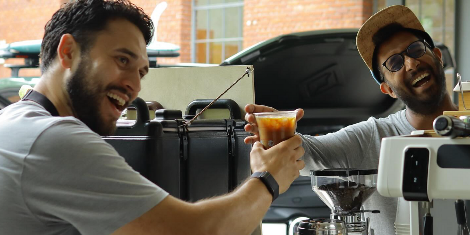 A barista hands an iced coffee to a smiling customer over the coffee bar at an outdoor cafe.