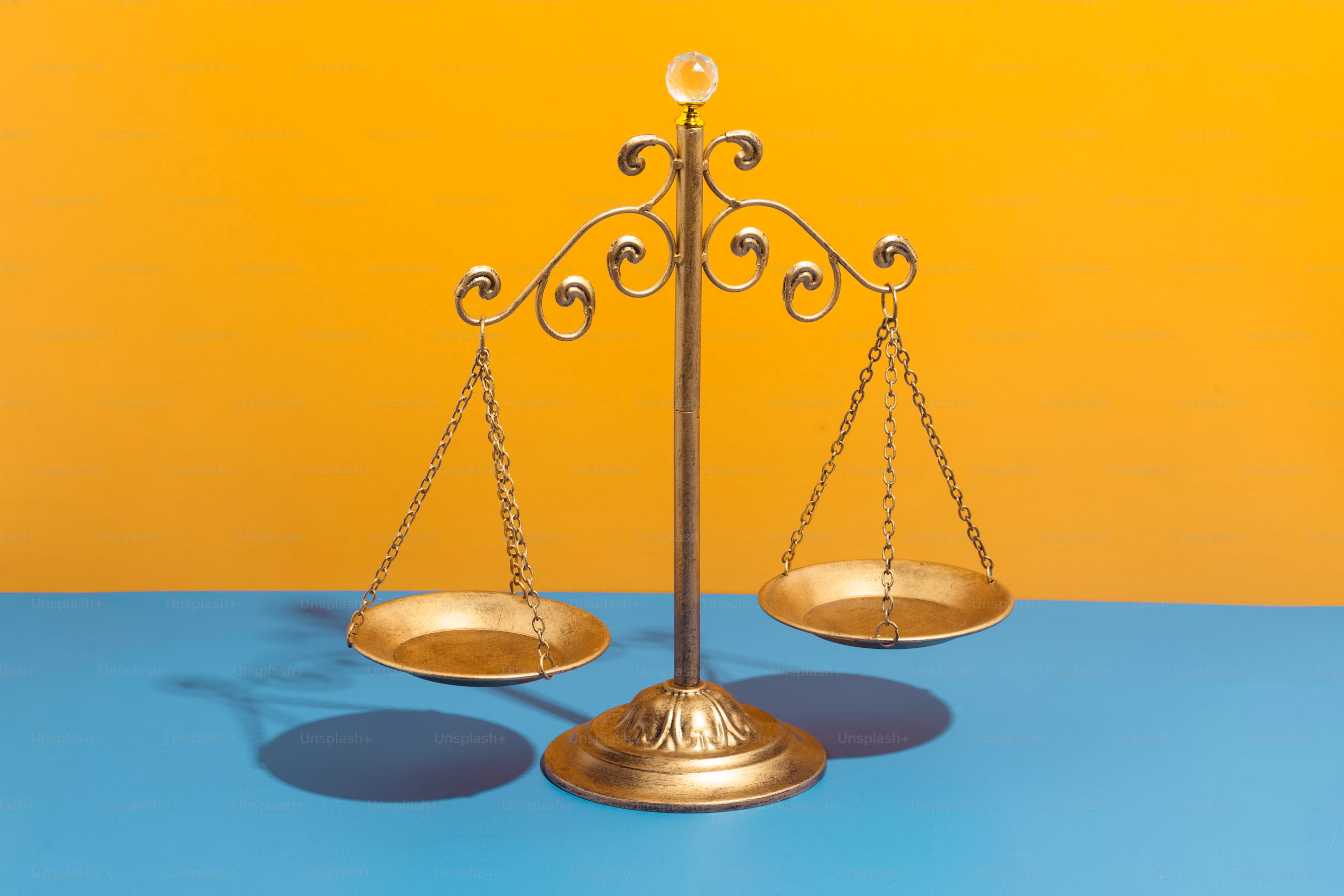 A gold variation on the scales of Justice sitting atop a blue surface on a yellow background.