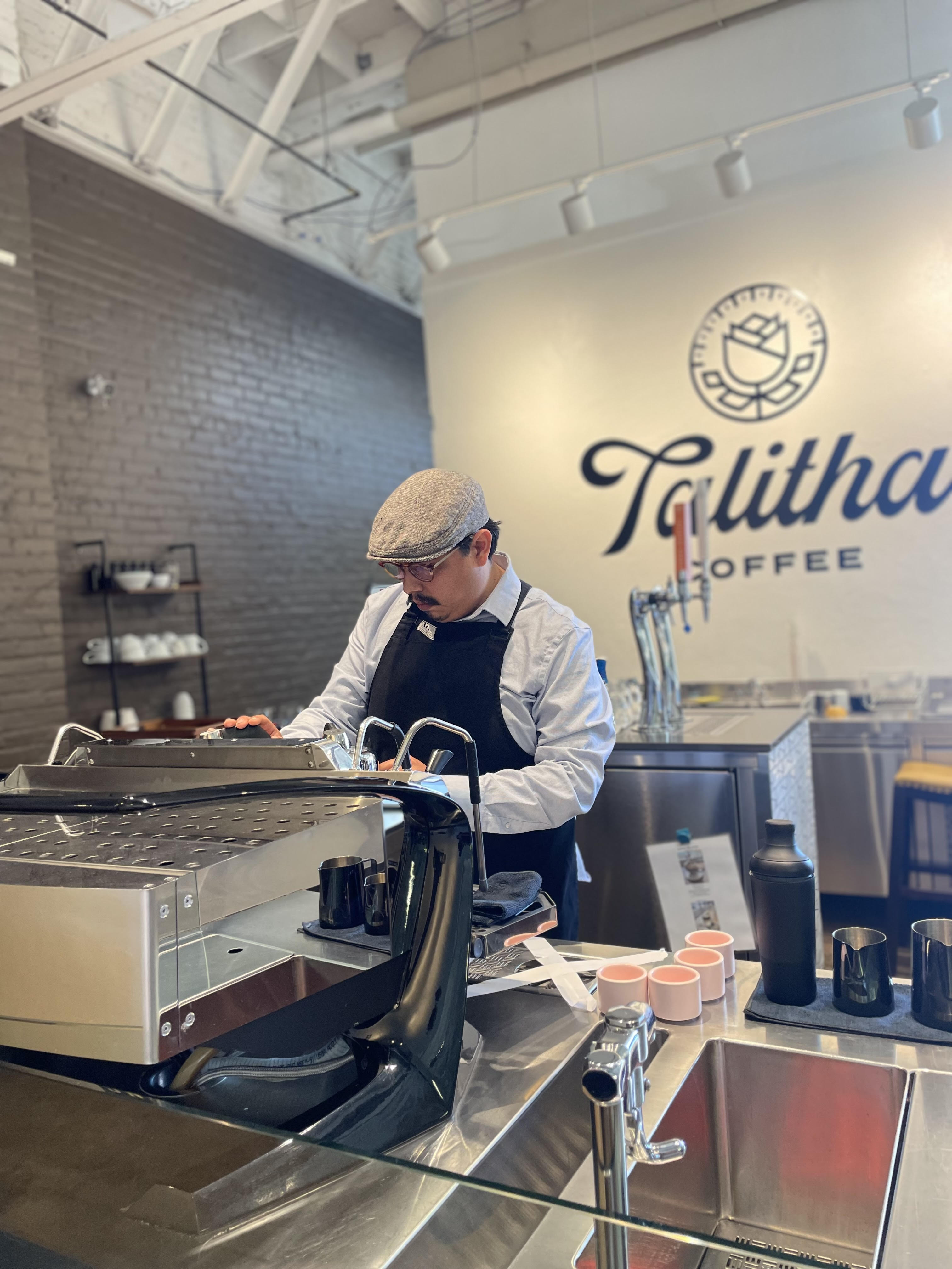 A mustachioed man wearing glasses, a newsboys cap, light button down under a black apron focuses intently on an espresso machine on a countertop in front of him.