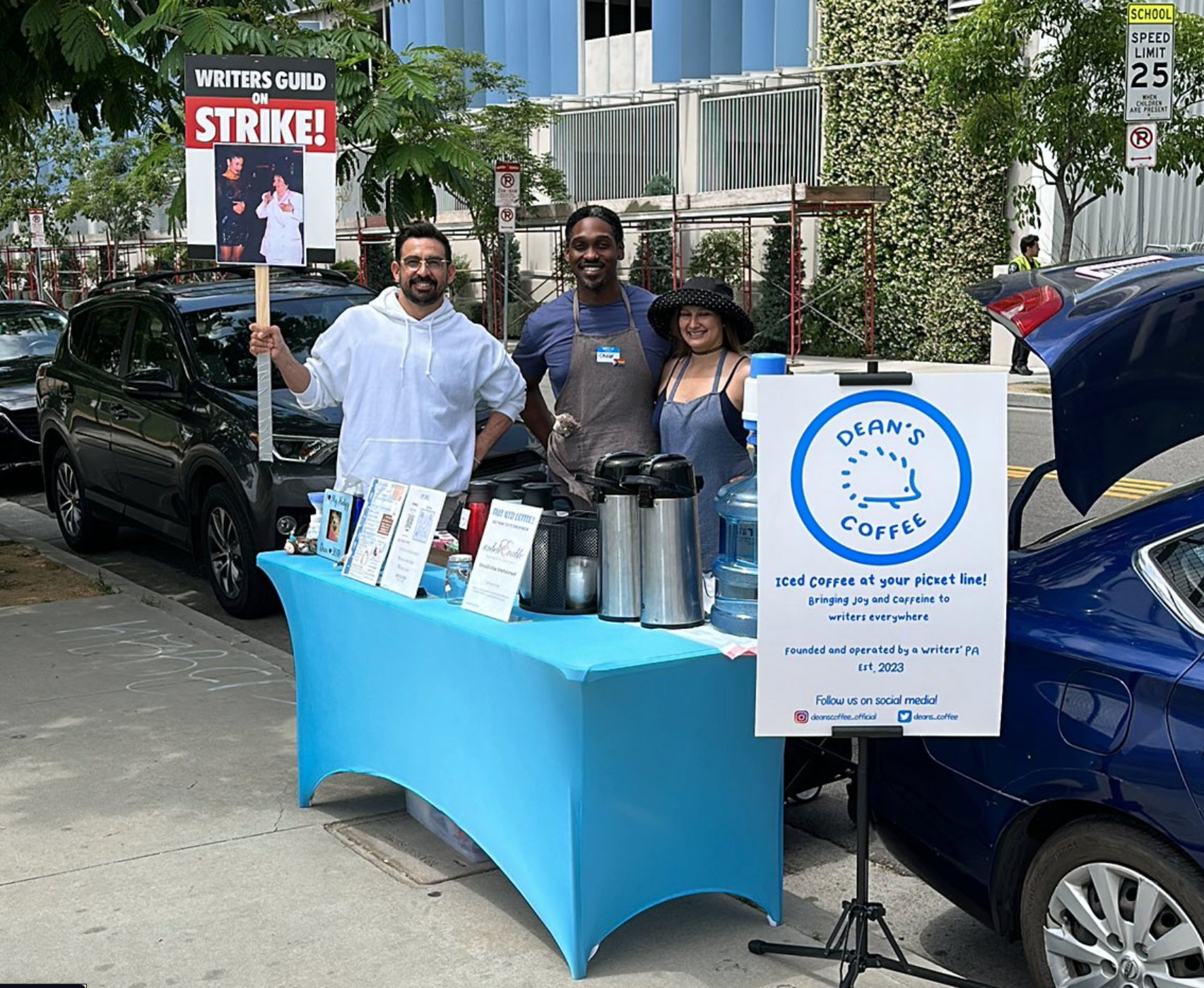 A picket line striker holding a Writer's Guild On Strike sign poses and smiles next to the Dean's Coffee pop-up table.