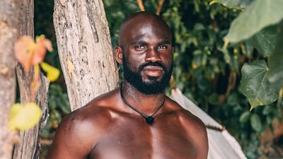 A man stares serenely down the camera lens as he stands between trees on an island