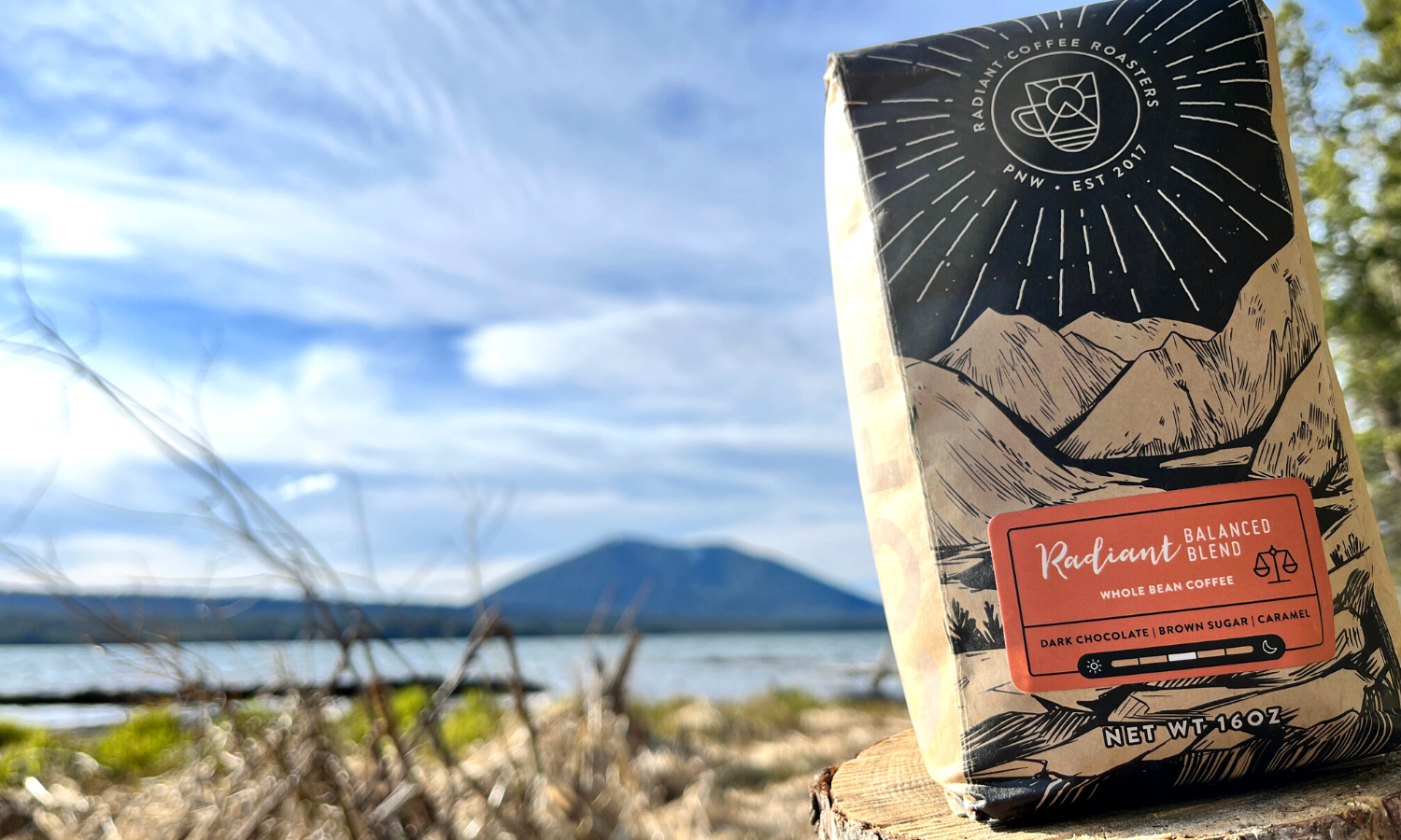 A blurred mountain against a blue sky brushed with white clouds  in the background. In the front, right, of the frame is a brown coffee bag with a mountainscape drawn across the front over the orange name label. 