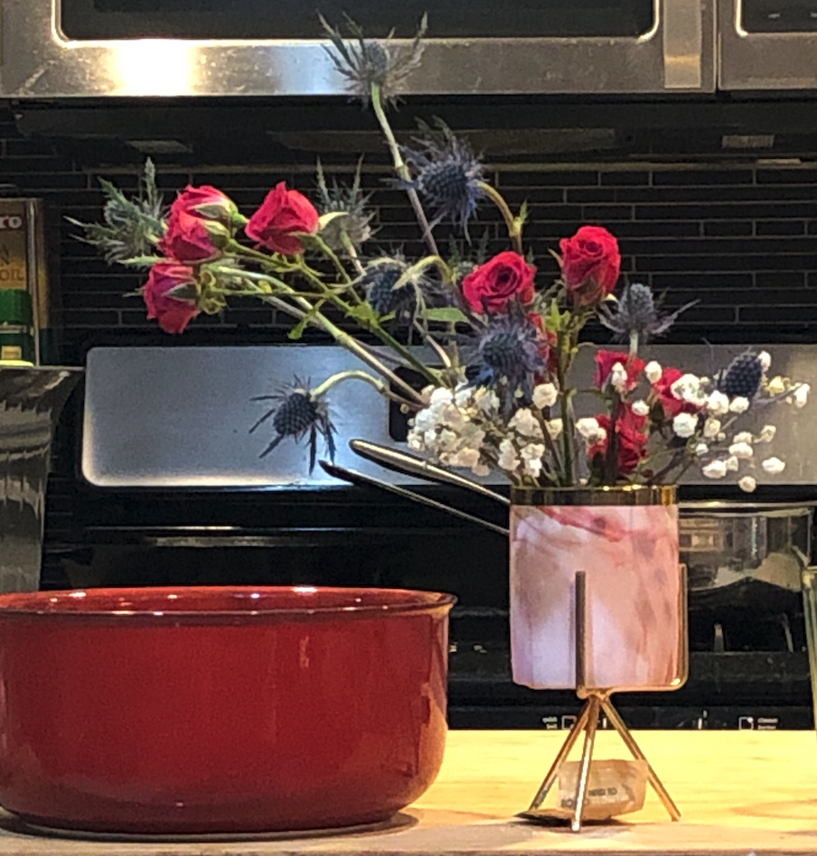 A bouquet of red roses, blue thistles, and white baby's breath in a small pink vase. You can also see a red bowl and an oven in the background.
