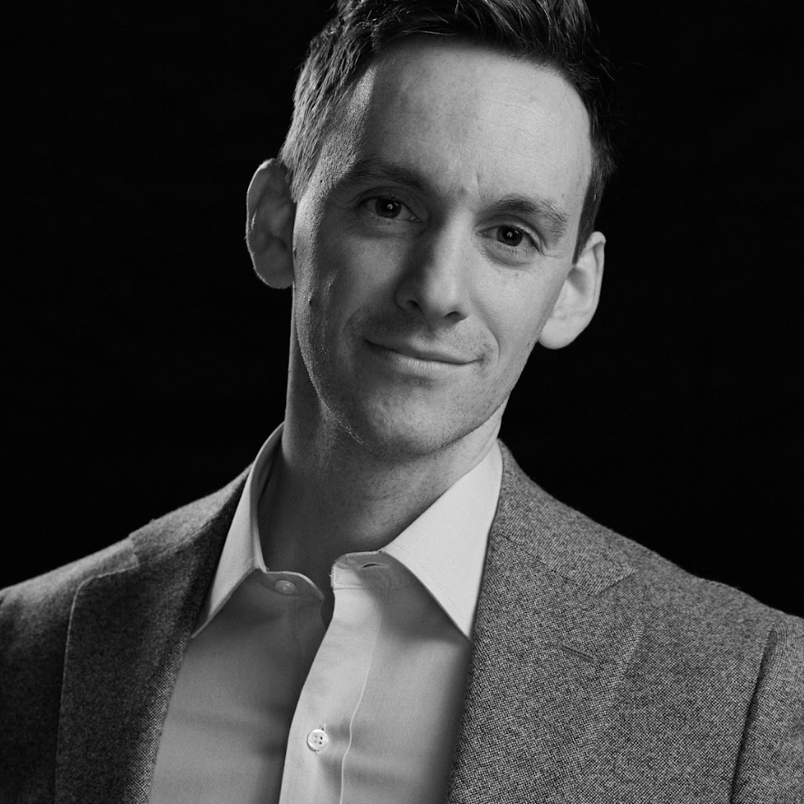 A black-and-white portrait of a man with short hair, wearing a light-colored button-up shirt and a textured blazer. He is facing the camera with a slight smile and a relaxed expression. The background is dark, which highlights his face and upper torso in the foreground.