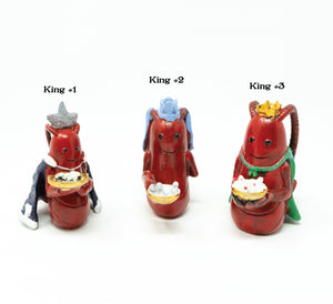 three small lobster replicas representing the three wise men wearing hats and capes and holding bowls of offerings in their claws labeled King #1, King #2, and King #3. King 1 is holding pearls, King 2 is holding baby seahorses, and King 3 is holding white star cookies with red centers