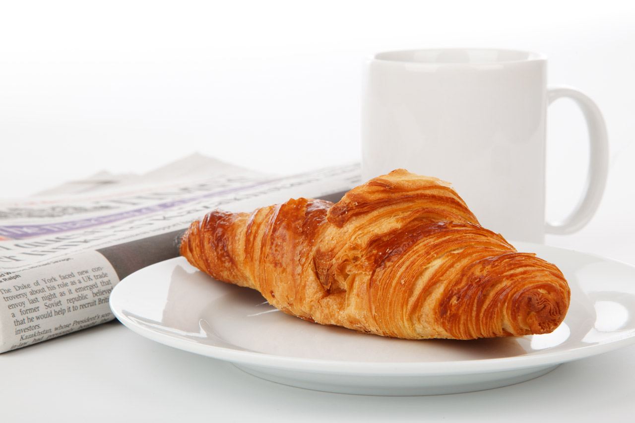 Golden croissant on a white plate beside a coffee cup and newspaper