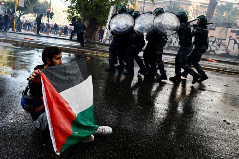 An unmasked person sits crosslegged on a road while holding up a Palestine flag with both hands. Riot police approach them with shields up An unmasked person sits crosslegged on a road while holding up a Palestine flag with both hands. Riot police approach them with shields up