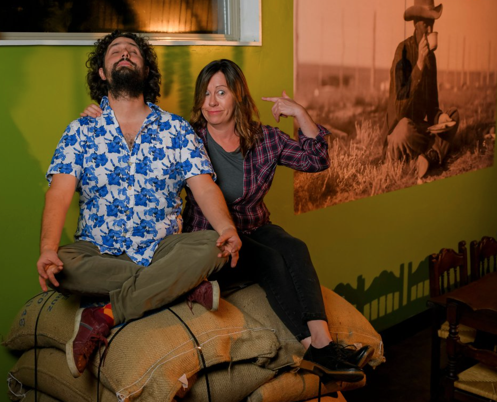 Weston and Sam Nawrocki making a funny face sitting atop a stack of coffee beans in burlap sacks in front of a green wall.