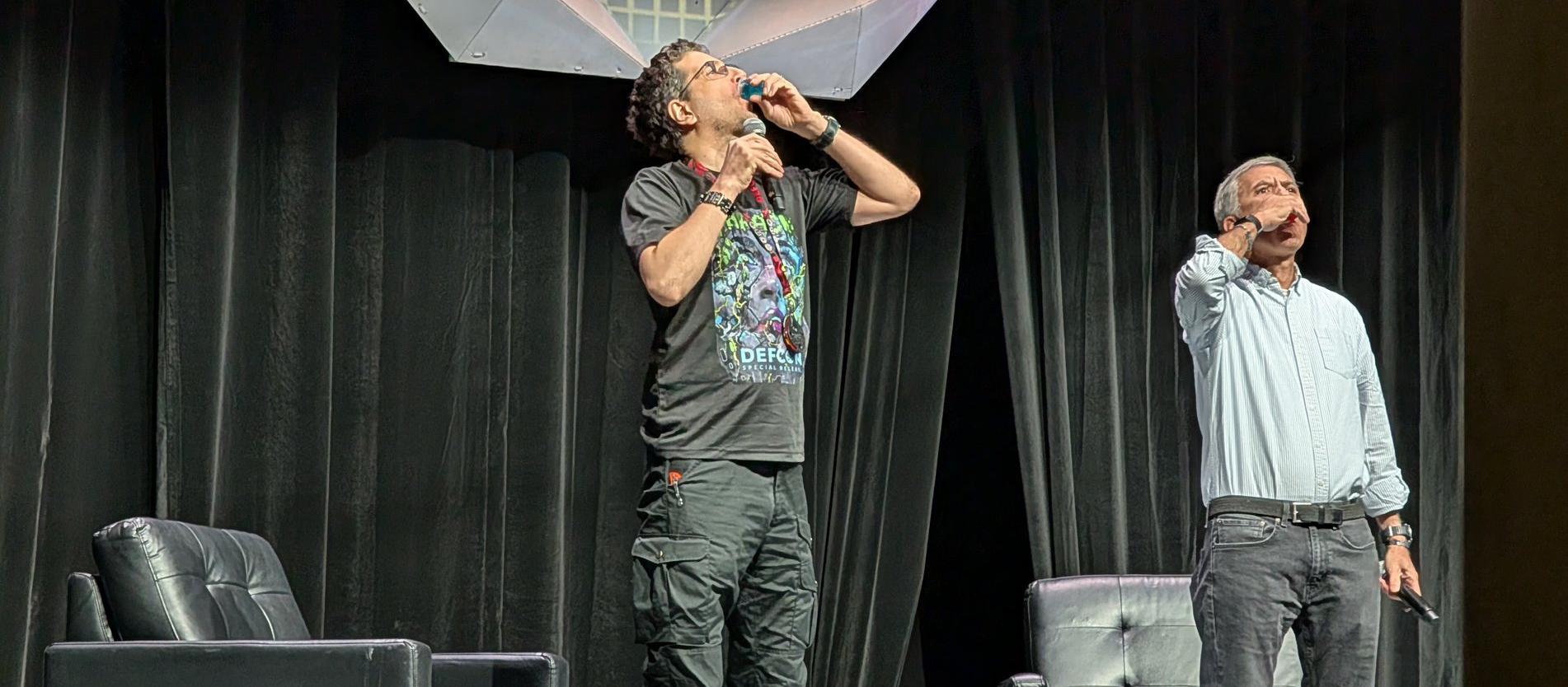 The image shows two men standing on a stage with black curtains in the background. Both men are taking a shot from small glasses, each holding their glass to their mouths in mid-drink. The man on the left has curly hair, wears glasses, a black graphic t-shirt, and cargo pants, while the man on the right has short gray hair, wears a light blue button-up shirt and jeans. There are two black leather chairs positioned on the stage, one near each man.