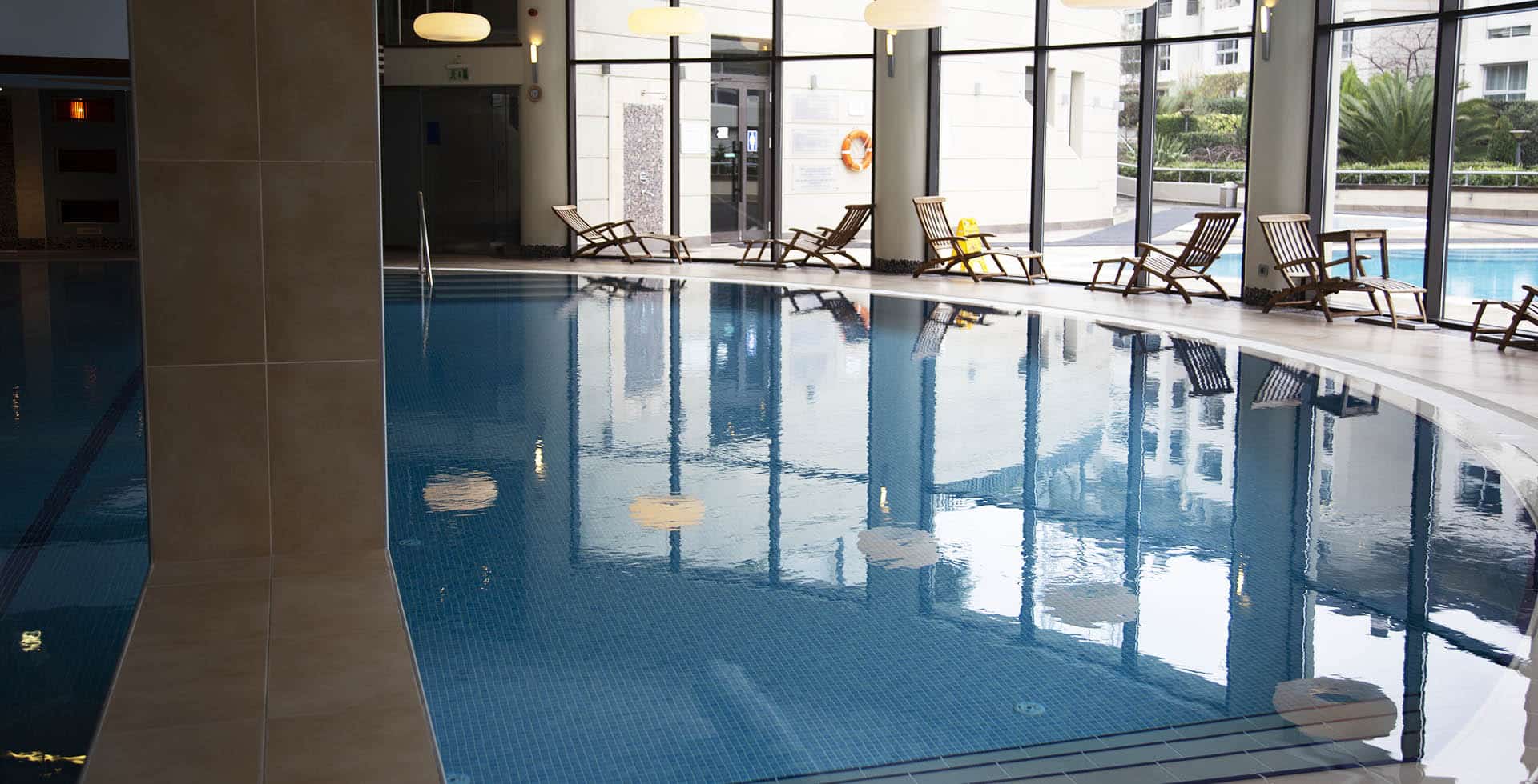 Curved indoor pool in a residential project in Istanbul: blue mosaic lining, a pale stone deck with timber loungers along a glazed wall and an outdoor pool visible beyond the glass
