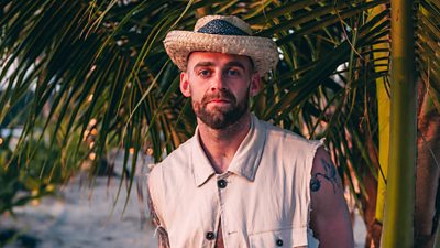 A man in a straw hat and sleeveless shirt stands against a palm tree on a beach