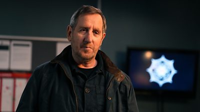 A man in a dark jumper and green jacket stands in a police station smiling at someone off camera