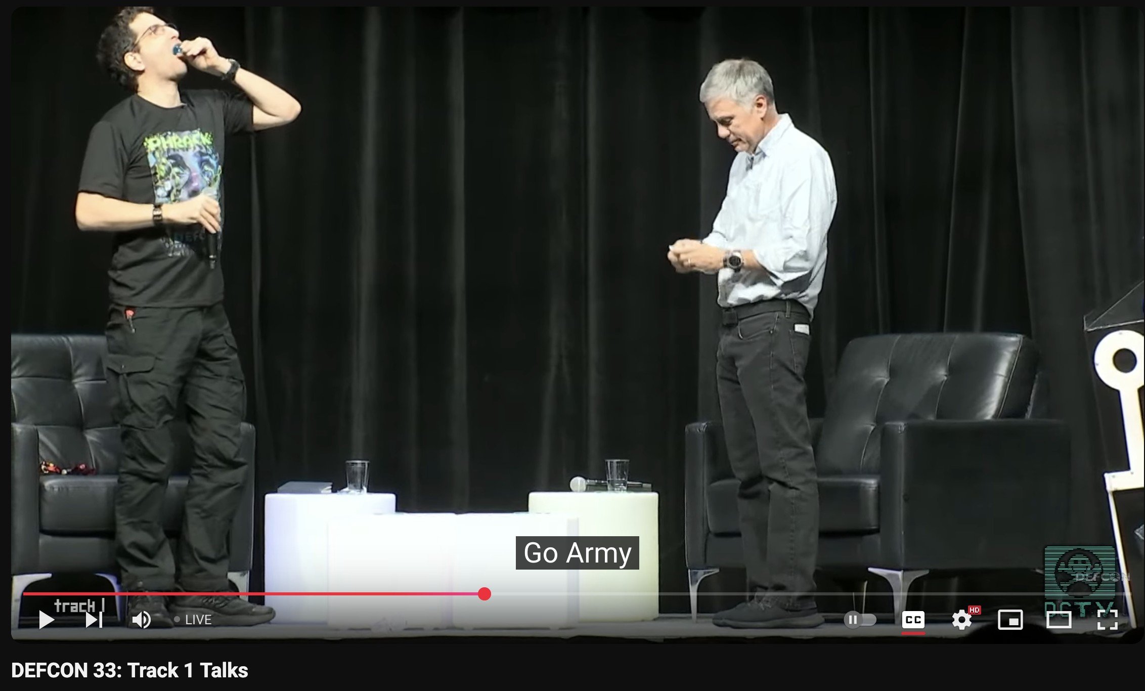 The image shows two men on a stage with a black curtain backdrop, engaged in an activity. The man on the left, wearing a black graphic t-shirt and black cargo pants, is leaning his head back and appears to be swallowing or placing something in his mouth while holding a microphone in his other hand. The man on the right, wearing a light blue button-up shirt and dark pants, is looking down and focused on an object in his hands. Two black armchairs and a white table with glasses and a microphone are positioned between them. The screen caption reads "Go Army," and the event is labeled as "DEFCON 33: Track 1 Talks."