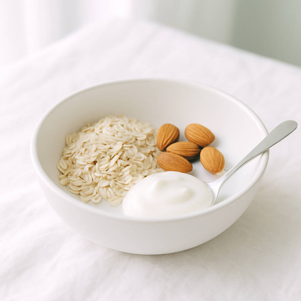 Light and airy close-up of a white ceramic bowl filled with plain whole-food ingredients — a small pile of rolled oats, a few whole almonds, a spoonful of natural yoghurt — on a bright white linen cloth in natural daylight, no packaging, no labels, soft sage green accent