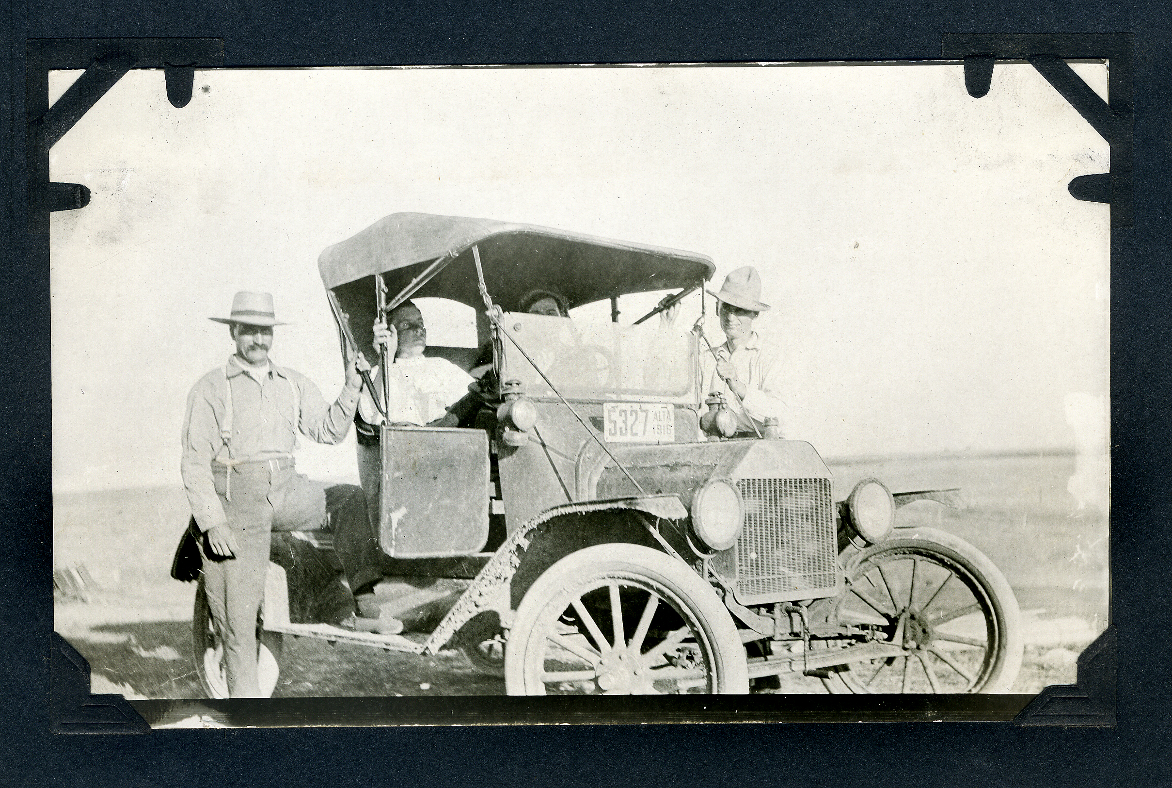 The image shows an early 20th-century automobile with four people either sitting in or standing beside it. The car has large, spoked wheels and a soft canopy roof. Two men stand outside the vehicle, one on each side, both wearing wide-brimmed hats and light-colored shirts with suspenders. Inside the car, two people are seated, one of whom appears to be the driver. The background is an open, flat landscape, suggesting a rural or undeveloped area. The photo has an aged, black-and-white appearance typical of early photography.