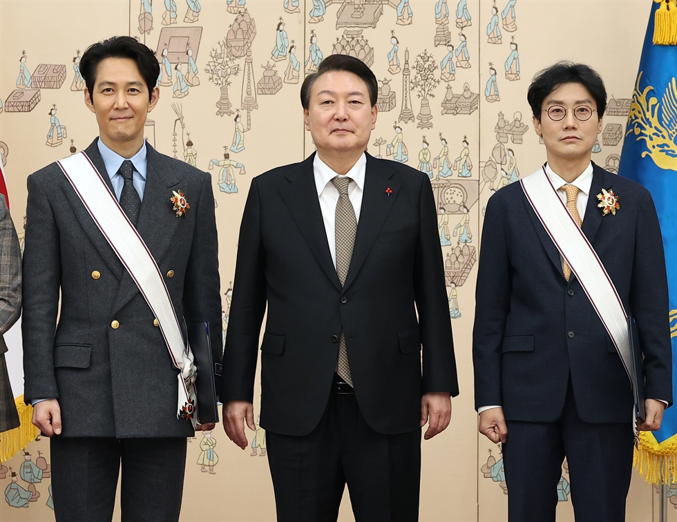 Kültür empozisyonunu ulusal bir mesele yapan Güney Kore (bakınız senelerdir yatırım yaptıkları edebiyat alanında bu yıl Han Kang ile Nobel ödülünü ülkeye getirmeleri veya K-Pop grubu BTS’in varlığı), Squid Game ’in Netflix’te en çok izlenmiş yapım olması ve kültüre katkılarından ötürü, dizinin yaratıcısı Hwang Dong-hyuk (sağda) ve başrol oyuncusu Lee Jung-jae’yi (solda) 2022’de Altın Taç Kültürel Liyakat Nişanı ile onurlandırmıştı. Ortada, nişanı takdim eden azledilmiş başkan Yoon Suk Yeol.