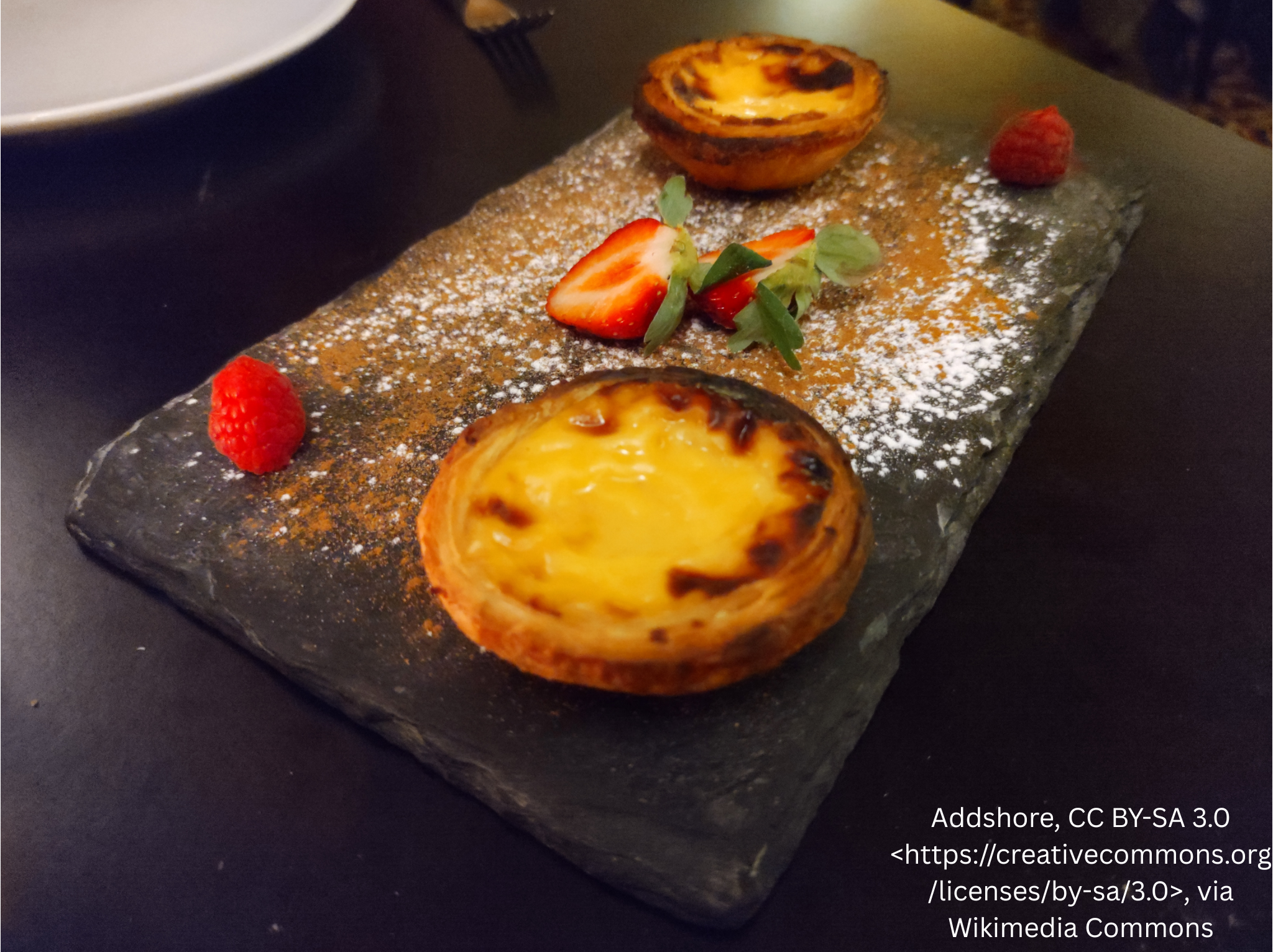 Two pastel de nata tarts with strawberries on a dark serving plate