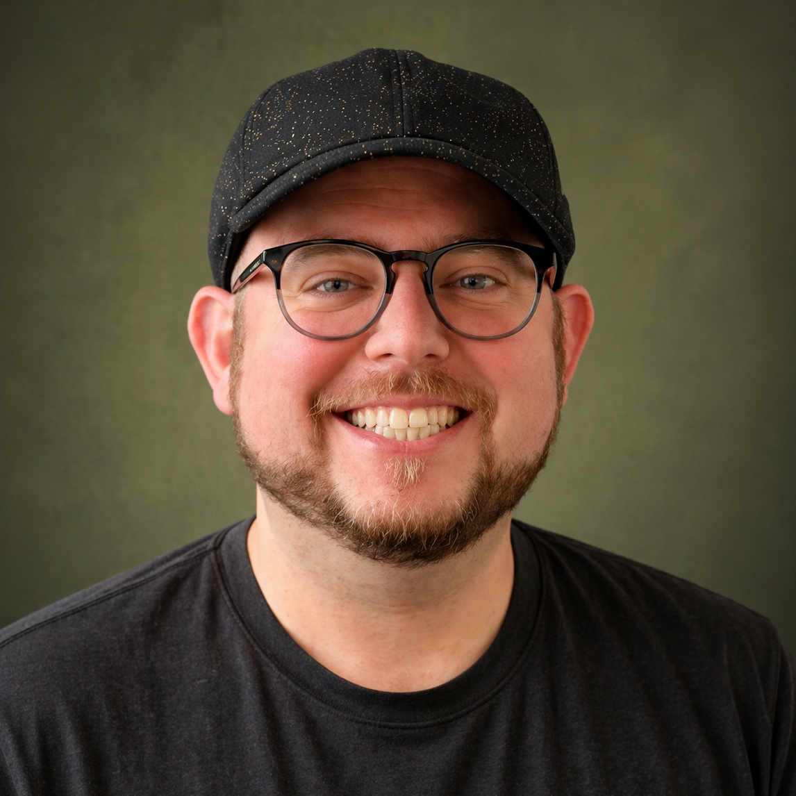 A smiling man in a dark cap and glasses with a beard, standing in front of a green backdrop