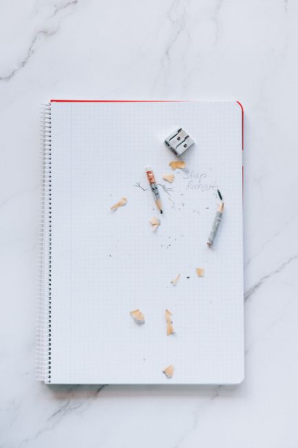 Overhead flat lay of a small notepad with a pen beside it, a glass of water, and a few green leaves on a white marble surface, bright natural daylight, clean and minimal