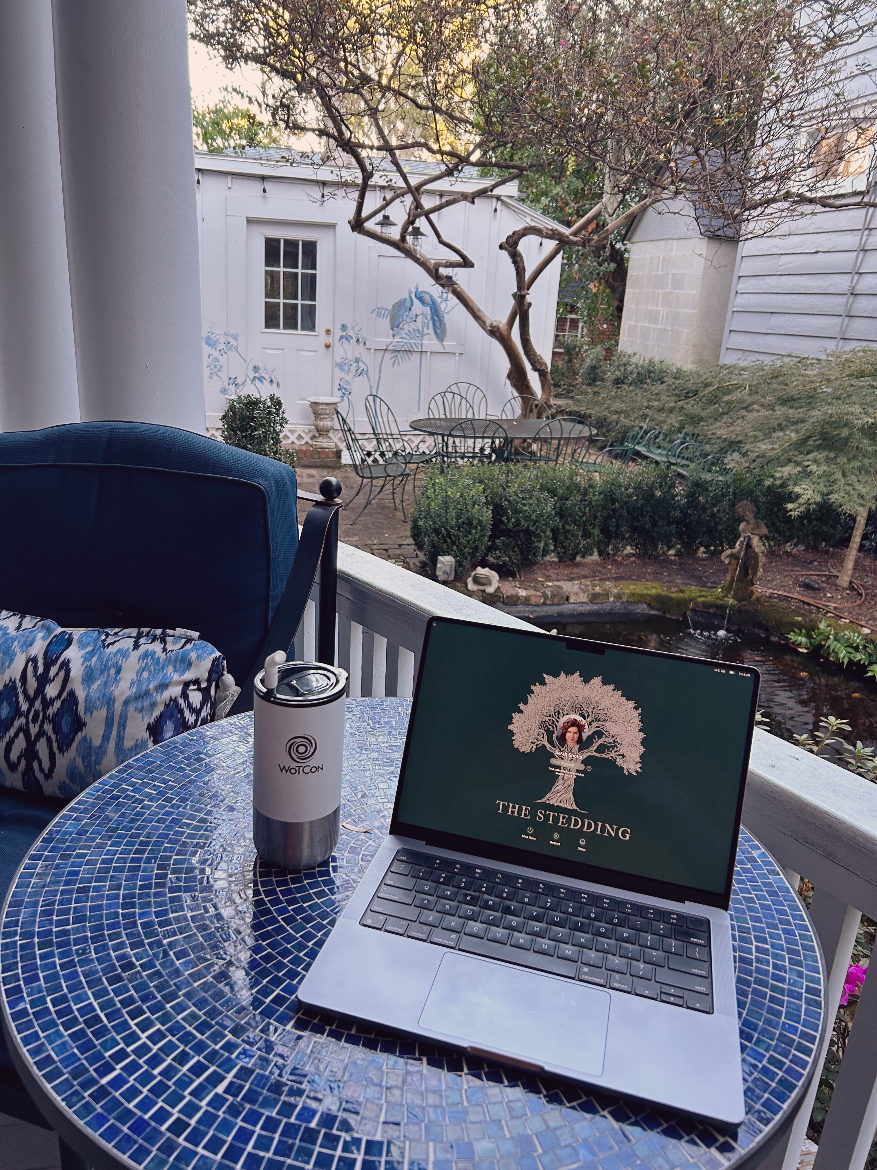 Laptop with The Stedding wallpaper and WoTCon wallpaper tumbler on a blue mosaic table on porch. Garden with tree, pond, and shed with hand painted peacocks beyond.