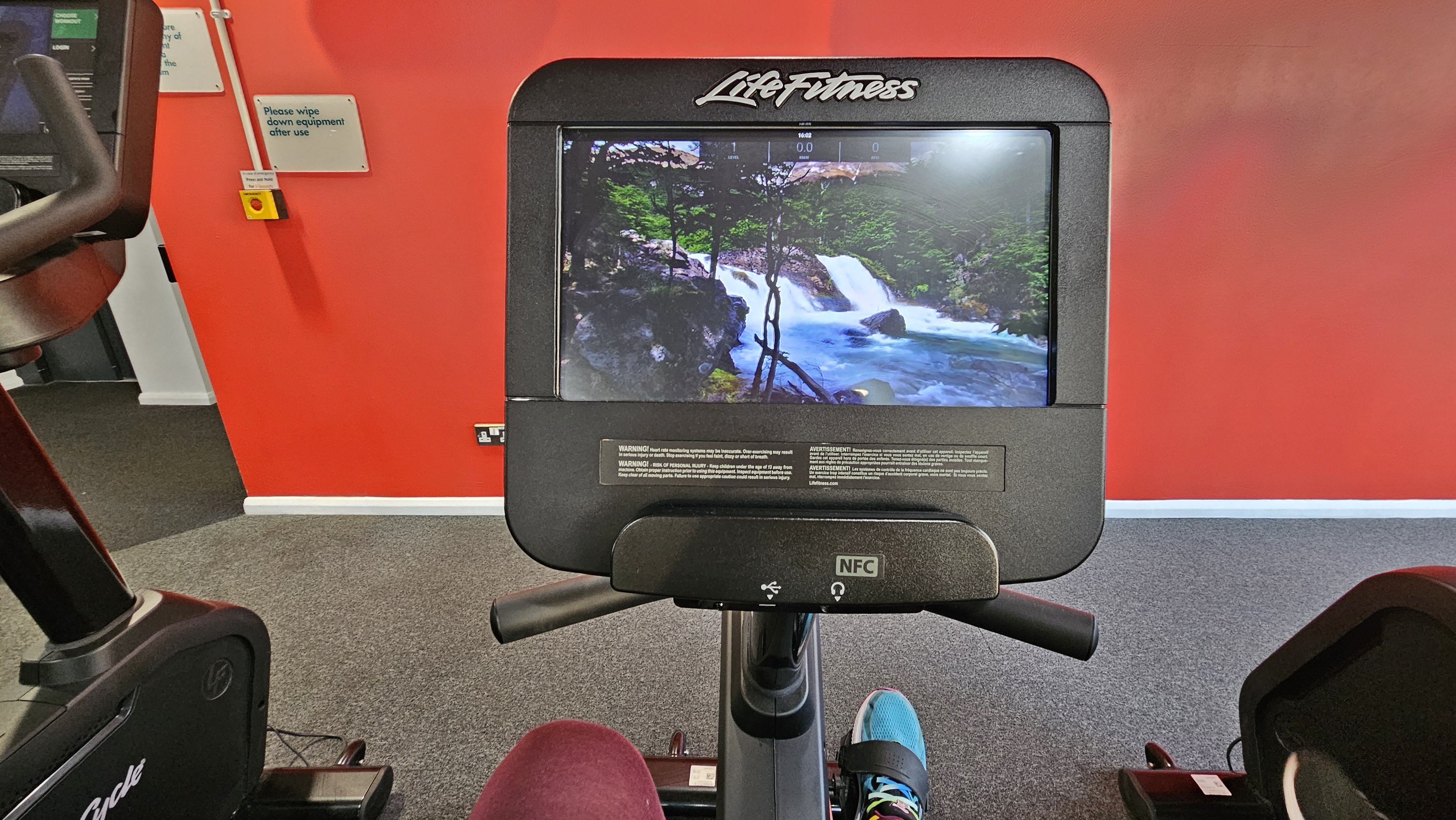 POV desde una bici estacionaria en el gimnasio. La pantalla de la bici muestra un video de unas cascadas en la Patagonia.