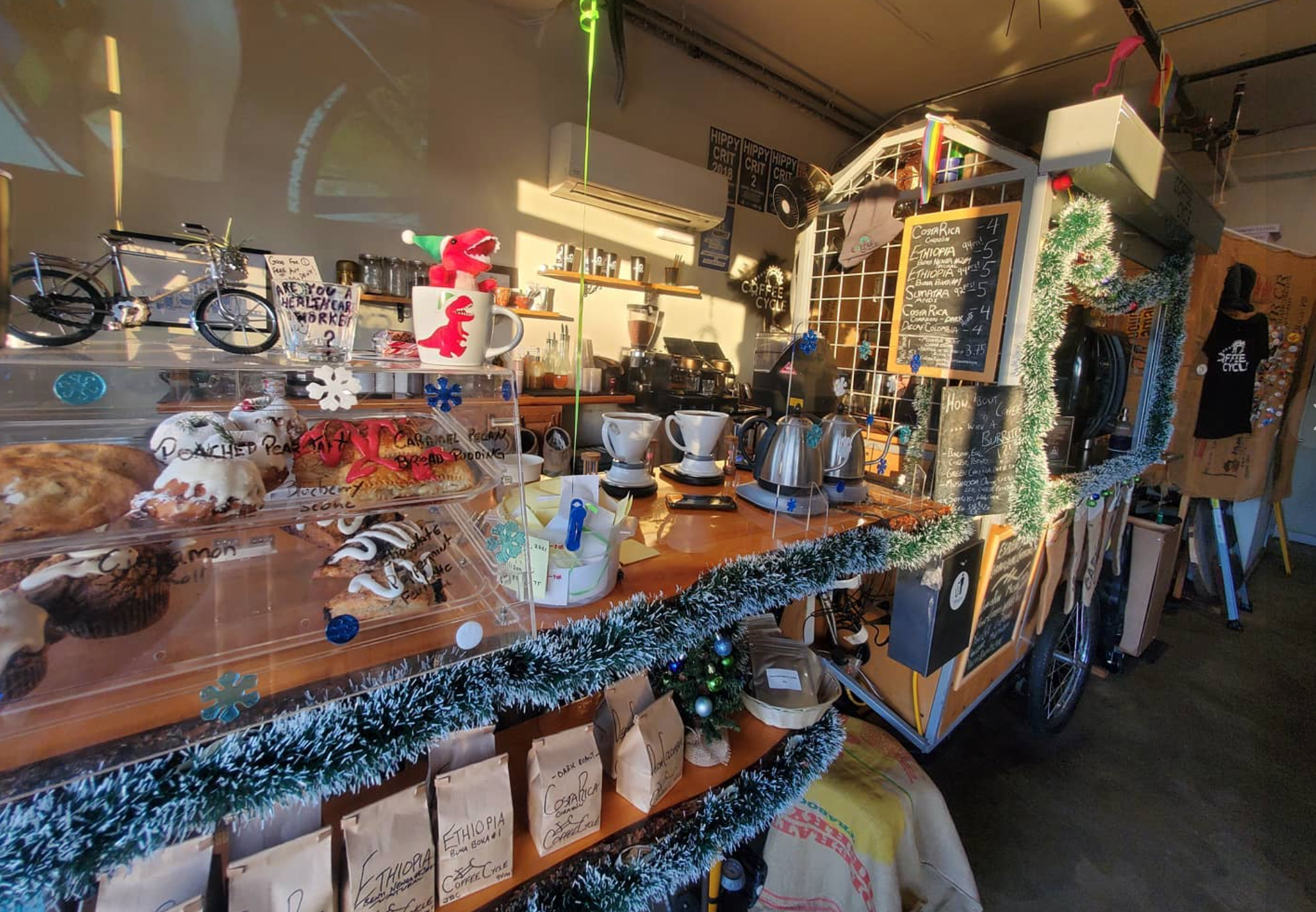 A long shot from left to right of the coffee bar at Coffee Cycle cafe in Pacific Beach San Diego. Wood bar top with coffee kettles, a pastry card, a dinosaur christmas decoration and on a shelf below brown bags of coffee beans for sale with handwritten labels.