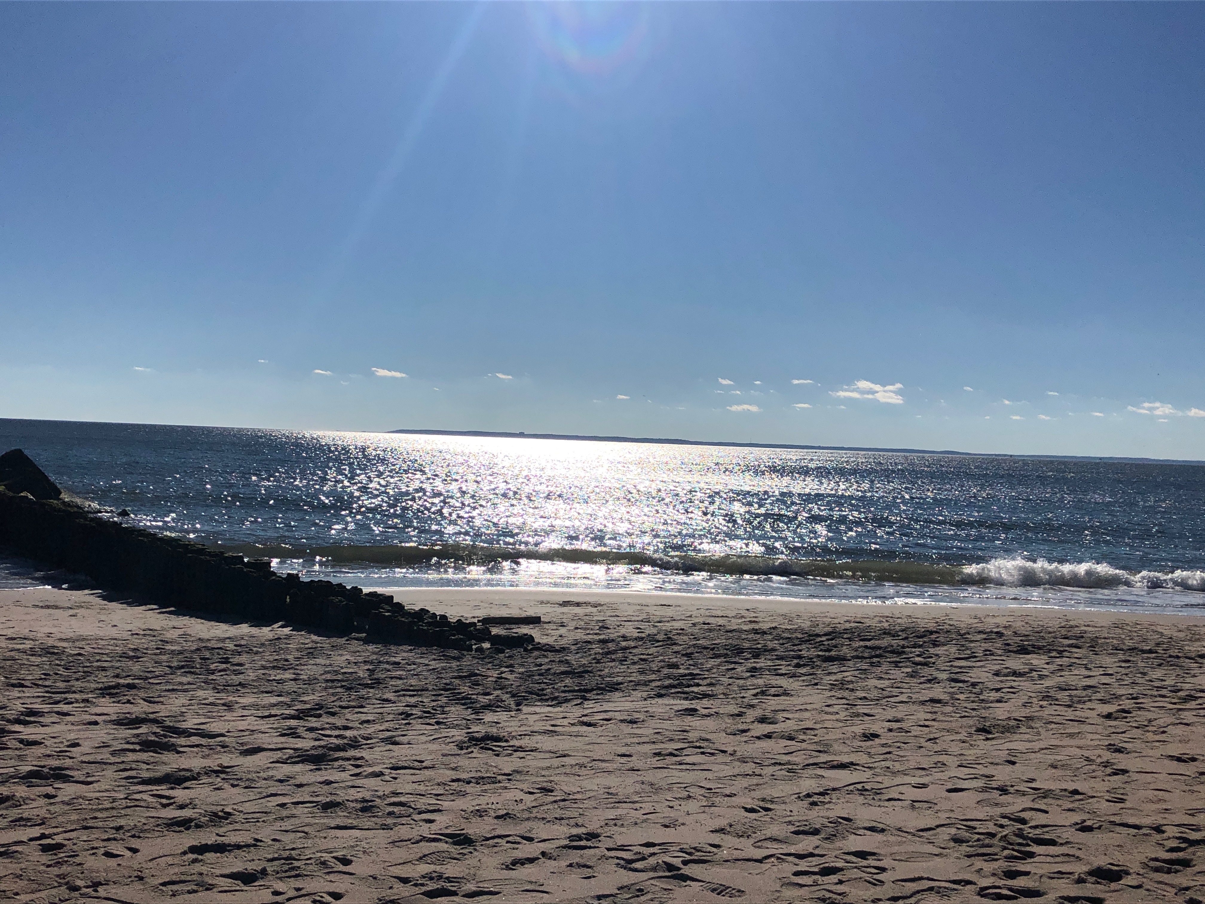 The beach and the ocean, with a rocky breakwater on the left. The sun is shining on the water.