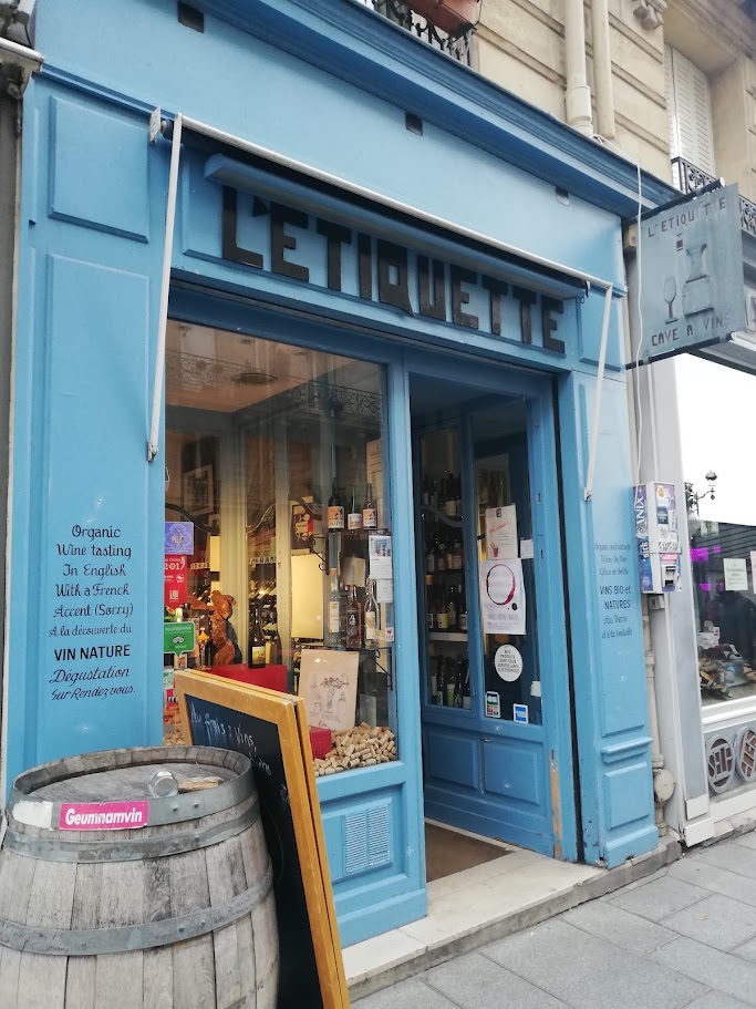 A photo of L'Etiquette. A blue shop with wine bottle sin the window. A sign reads "Organic wine tasting in English with a French accent (sorry)