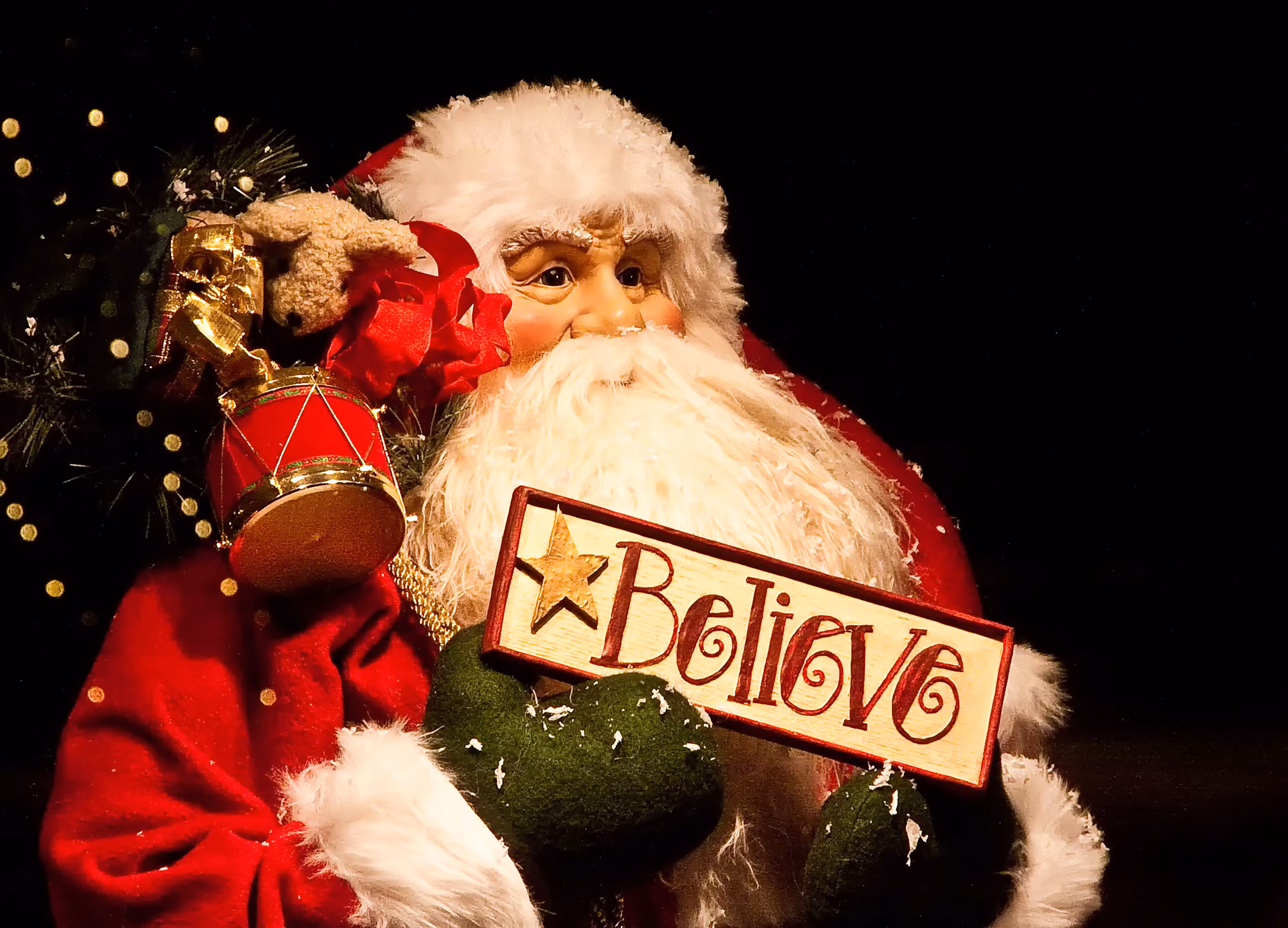 A picture of a Santa Claus figurine, probably about two feet tall though it's impossible to tell precisely from the angle the picture was taken from. The background is black, and there's the tip of a branch of what is probably a Christmas tree in the background, with blurred lights and a few visible needles. The branch extends over Santa's right shoulder, and there's a red and gold drum ornament hanging down onto his shoulder. Santa himself wears the traditional red coat and hat with white faux fur trim, has a big white bushy beard, and features dark, almost creepily realistic eyes with crow's feet wrinkles that make them look happy. His cheeks are very rosy. Against his chest, in hands with green mittens, Santa holds up a rectangular sign--white with a red border--on which there's a yellow stare and the word "believe" written in red.