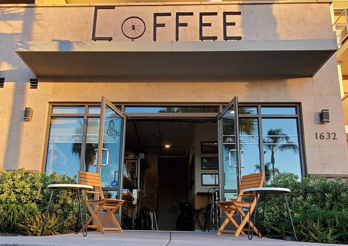 A photo from a low angle of the entry way to Coffee Cycle cafe in Pacific Beach, San Diego, California. A chair and side table frame the open doorways and the word coffee in black iron wrought letters is mounted above the door.