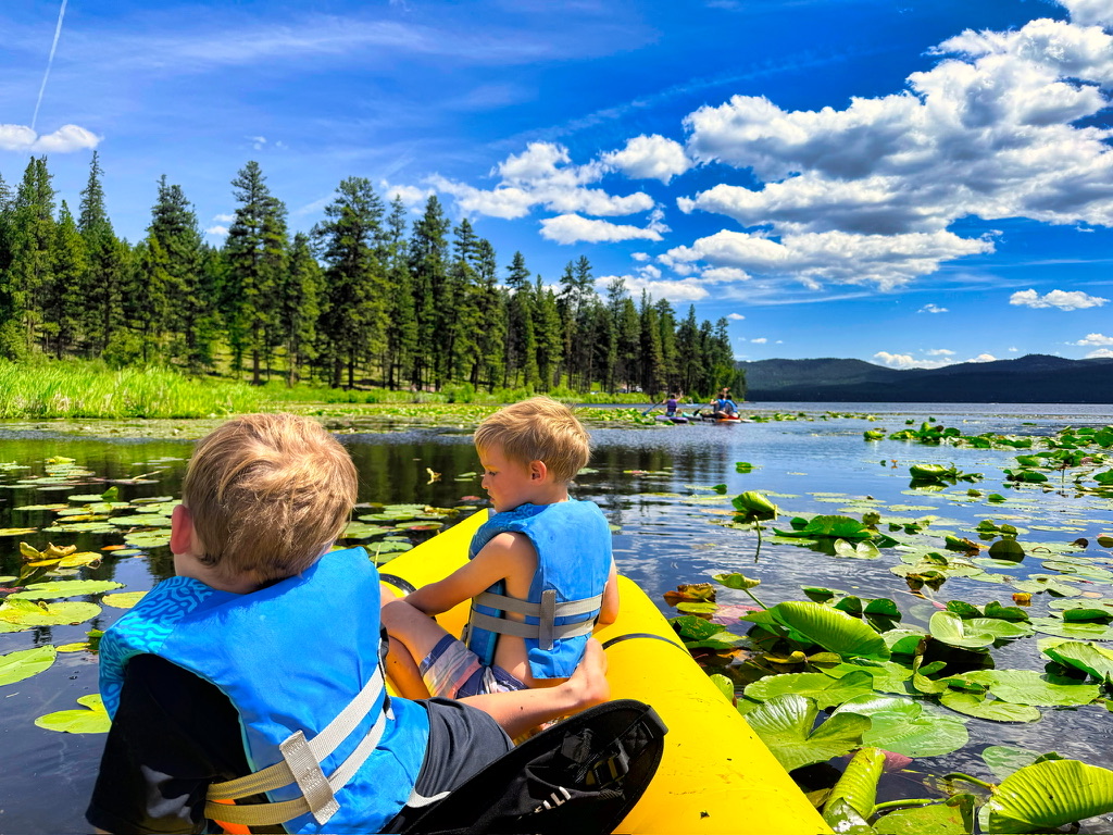 Two children wearing blue life jackets in a yellow kayak among lily pads on a lake.