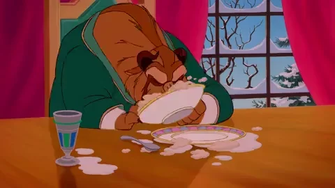 A GIF of the Beast from Beauty and the Beast. He is wearing a gran coat, sitting at a table, eating a bowl of food by shoving his face into it.