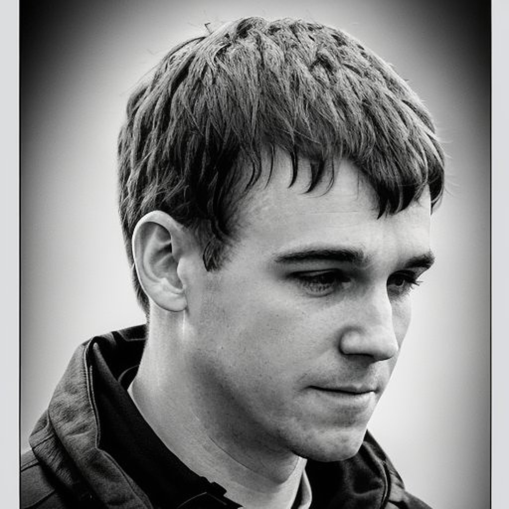 The image is a black and white close-up portrait of a young man with short, slightly tousled hair. He is looking downward and to the side with a calm, contemplative expression on his face. He wears a dark jacket over a collared shirt, and the background is softly blurred, emphasizing his facial features.