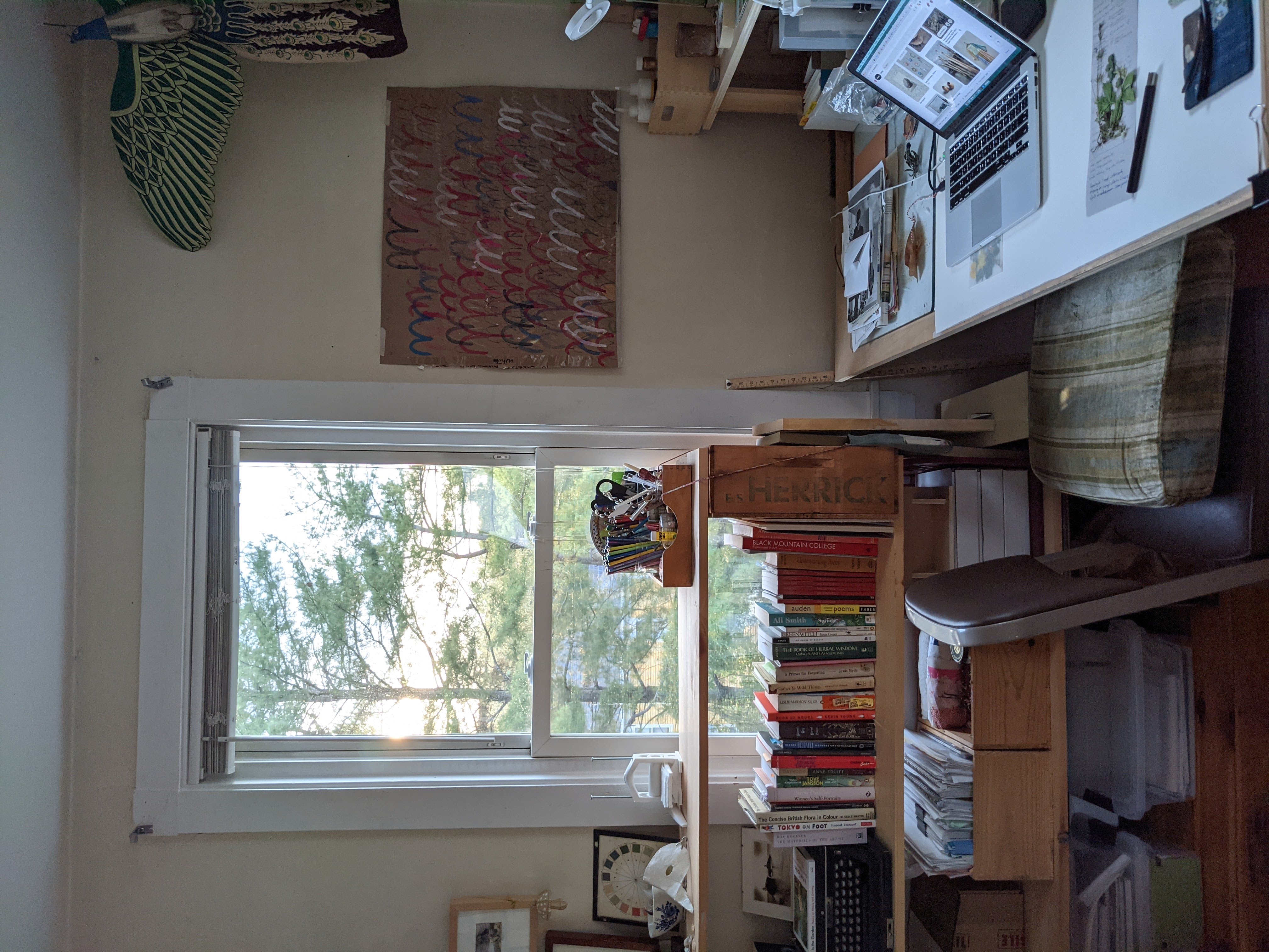 a room with bookshelves made of boards and boxes, a desk made of a board and trestles, a chair. books, pictures on the wall, a kite shaped like a peacock.