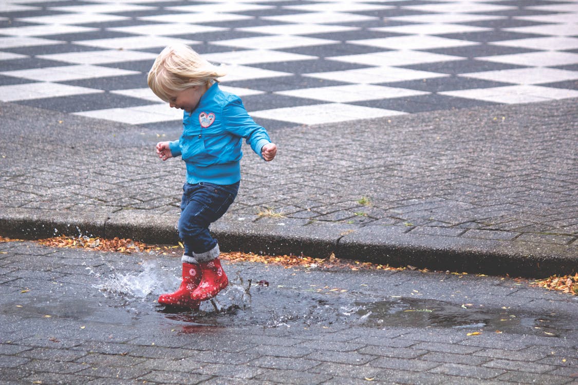 Free A young child joyfully splashes in a puddle wearing red boots on a rainy day. Stock Photo