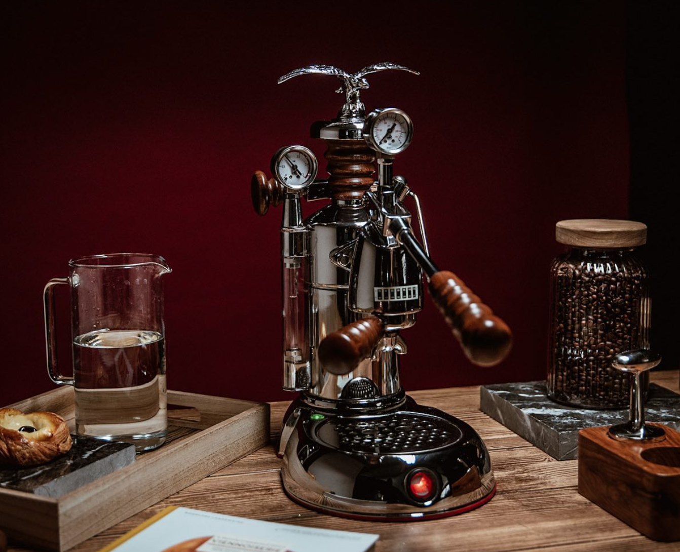 A shiny silver espresso machine that looks very complex with wooden handles and an eagle statue on top stands out against a purple-brown backdrop and is surrounded by various coffee accessories.