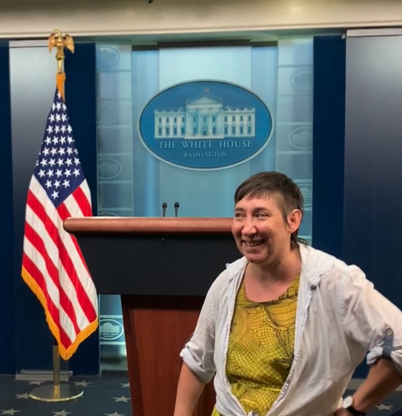 me, a white person, wearing a yellow dress and a blue button up shirt, in front of the white house press pool room