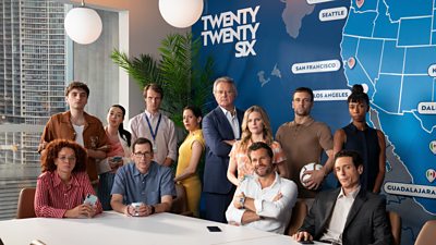 The cast of Twenty Twenty Six stand and sit around a large with a blue map on a wall behind them, featuring cities involved in the football tournament