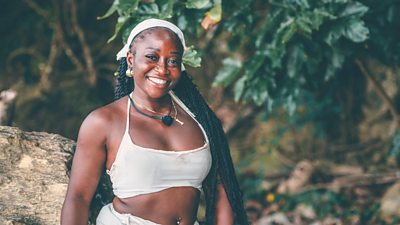 A woman in a white bikini top smiles to camera, standing against a backdrop of woods and green leaves