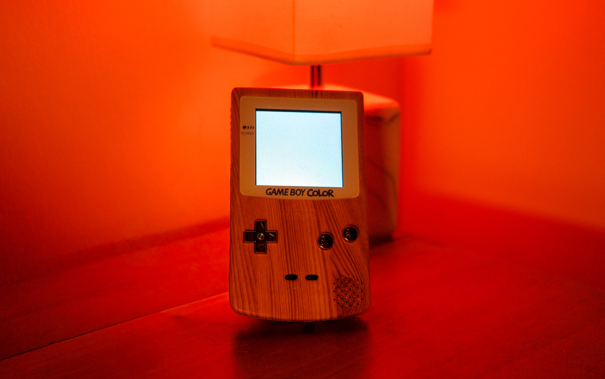 A photo of a modded Gameboy Color with a wooden shell sitting on a desk with a red light lamp behind it. The Game Boy screen is on, glowing white and blank.