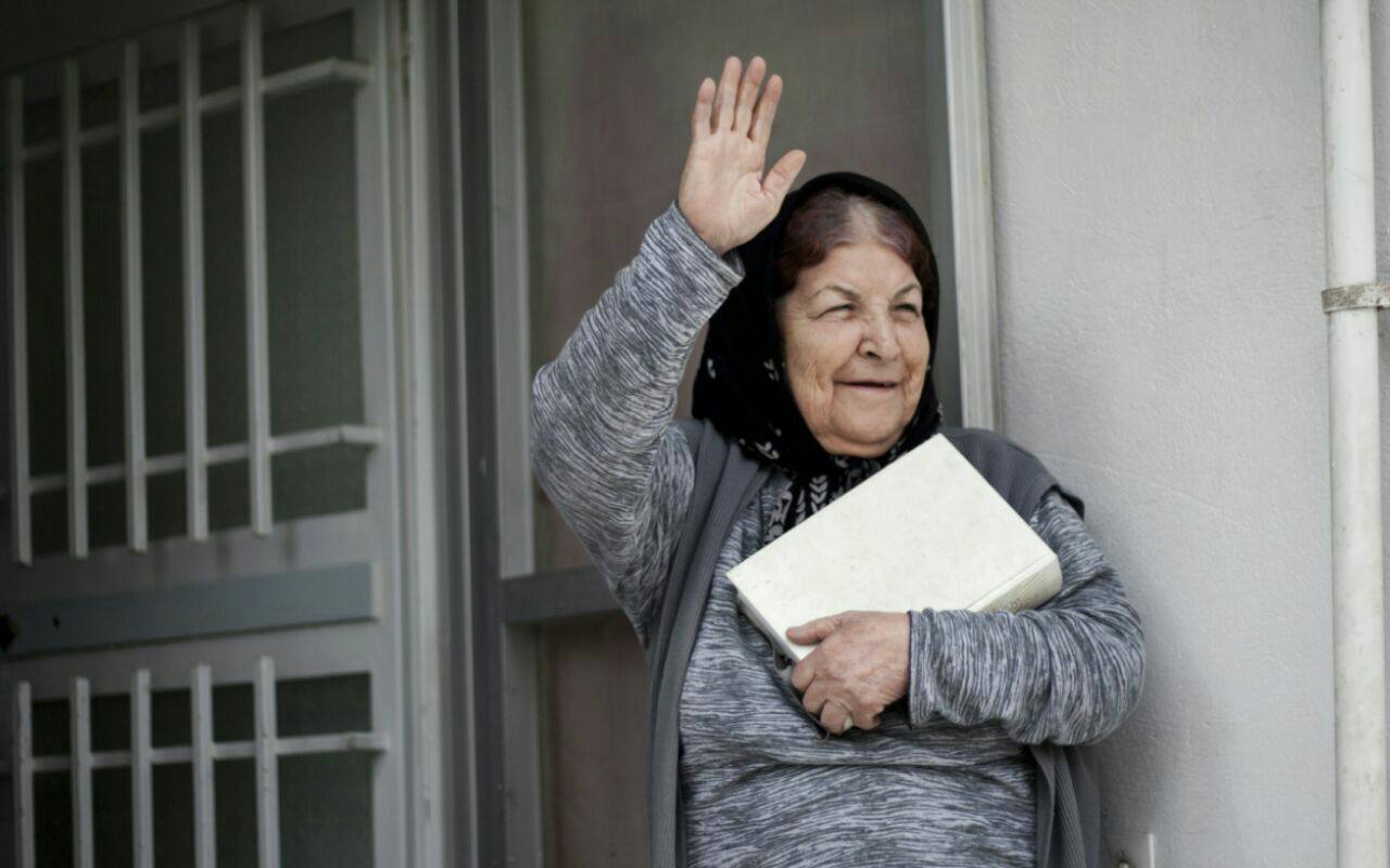 Older woman waving from a doorway while holding a folded item