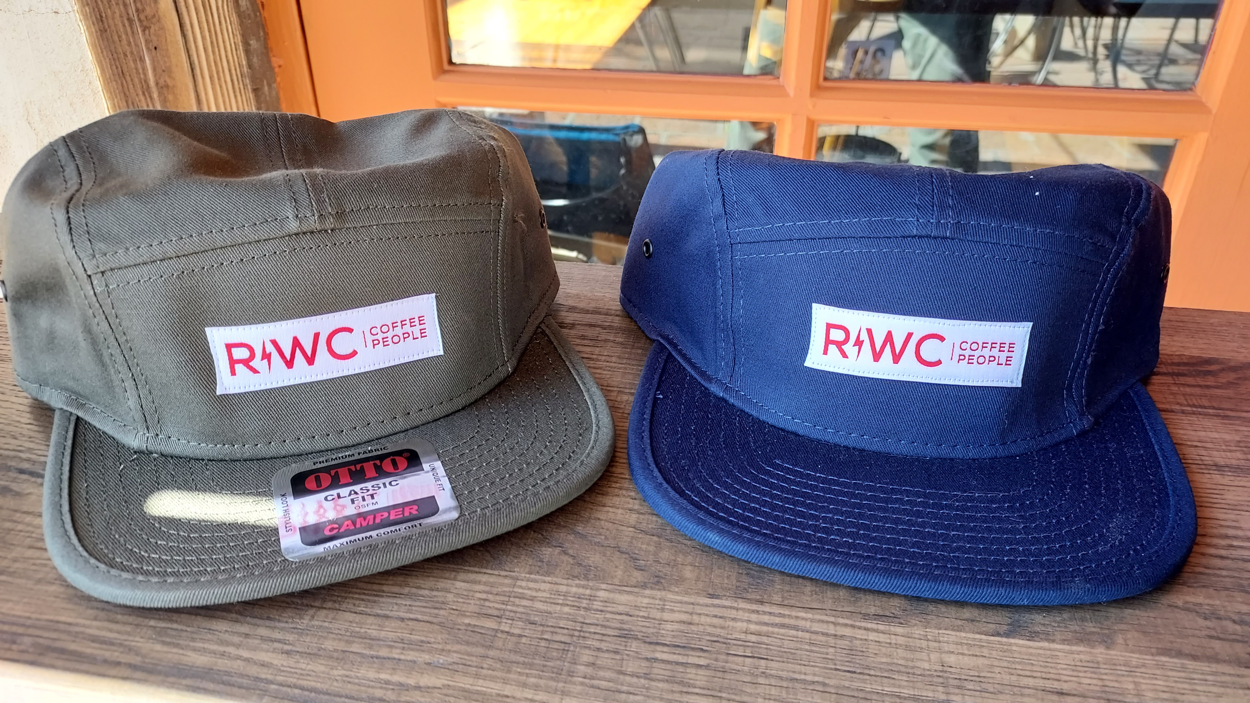 From Left: A grey 5-panel camp style cap with the R!WC Coffee People Logo and the same in nave sit on a wooden shelf.